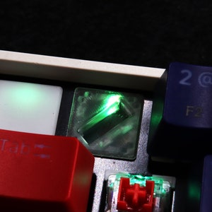 May include: Close-up of a mechanical keyboard with a green LED backlight. The Tab key is red, the 2 key is blue, and the F2 key is blue. The keyboard has a white frame.