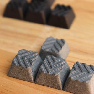 Peut inclure: Un ensemble de petites touches de clavier carrées présentant une surface texturée avec un motif en chevrons. Le lot comprend plusieurs pièces gris foncé, couleur charbon, et une pièce blanche, disposées en groupe sur une surface en bois clair.