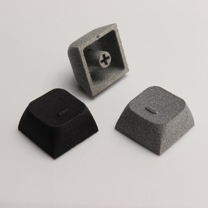 May include: Three keycaps in different colours: black, grey, and light grey. The keycaps are textured and have a square shape. The light grey keycap is shown from the bottom, revealing a screw hole.