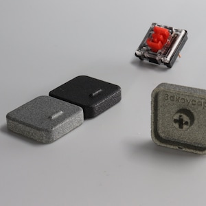 Puede incluir: Tres teclas diferentes para un teclado mecánico. Dos son grises y negras, y una es plateada con el texto "3dkeycap" y un signo más.