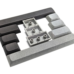 May include: A collection of mechanical keyboard keycaps featuring a mix of dark charcoal and light gray textured finishes. The set includes several rectangular and square keys, with three light gray keys shown from the underside to reveal the cross-shaped stems.