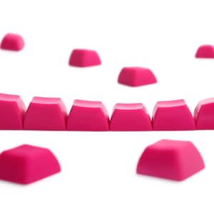 Könnte beinhalten: Eine Sammlung von zehn einzelnen, leuchtend pinkfarbenen Tastaturkappen, die in einer Reihe angeordnet und verstreut sind. Jede Tastenkappe hat eine glatte, matte Oberfläche mit einem leicht gewölbten Profil für mechanische Tastaturschalter.
