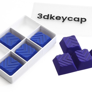 May include: A white divided container holding six blue keycaps with chevron patterns. Several additional keycaps are arranged nearby. A white card with the text "3dkeycap" is also present.