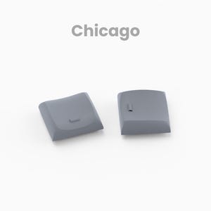 May include: Two gray, square-shaped keycaps with rounded edges are displayed on a white background. The word "Chicago" is written in gray above the keycaps. The word "Render" is written below the keycaps.
