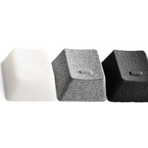 May include: Three mechanical keyboard keycaps arranged in a row. The first keycap is smooth and white, the second is a textured gray, and the third is a textured black. Each keycap features a small, horizontal dash symbol embossed on the front face.