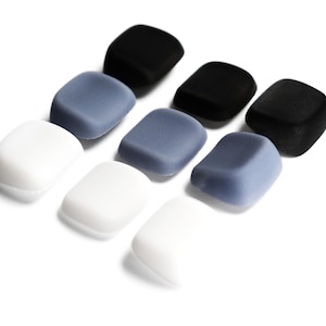 May include: A collection of eight small, smooth, square-shaped objects with rounded corners, arranged in a grid. The set includes three white, three slate blue, and two black pieces, all featuring a matte finish and a slightly concave top surface.