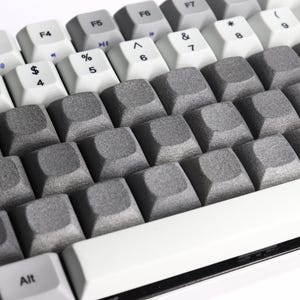 May include: A mechanical keyboard featuring a mix of white and light gray keys with standard alphanumeric labels and a row of function keys. The central section consists of blank, textured dark gray keycaps with a matte, granular finish.