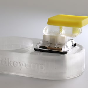 May include: A clear, 3D-printed keyboard keycap with a yellow key. The keycap is partially disassembled, showing the internal components. The base is clear with the text "3dkeycap" embossed on the side.