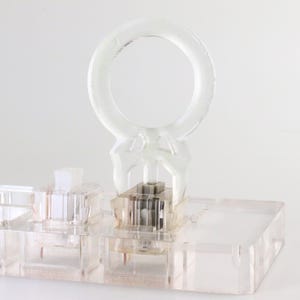 Könnte beinhalten: Ein transparenter Kunststoffring sitzt auf einer transparenten Kunststoffbasis mit mehreren Fächern. Der Ring ist mit einem transparenten Kunststoffmechanismus an der Basis befestigt.