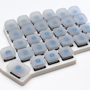 May include: A white keyboard with a grid of square, frosted-looking keys. Each key has a small, blue circular design in the center. The keyboard is on a white surface.