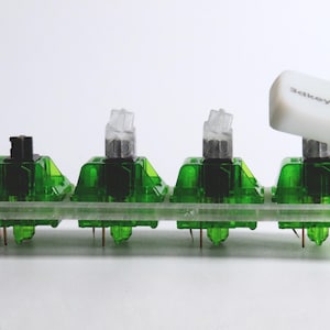 May include: A row of four mechanical keyboard switches. The switches have green housings and clear stems. One switch has a white keycap with the text "3akeycap". The switches are mounted on a clear plastic base.