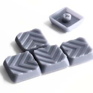 May include: A set of five square, light gray keycaps featuring a raised chevron pattern on the top surface. Each keycap has a smooth, matte finish and a cross-shaped stem on the underside for attachment to a mechanical keyboard switch.