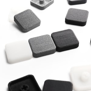Puede incluir: Una colección de pequeñas tapas cuadradas en negro, gris y blanco. Cada pieza presenta una superficie superior texturizada y un conector central en forma de cruz en la parte inferior, diseñado para fijarse a una base o componente.