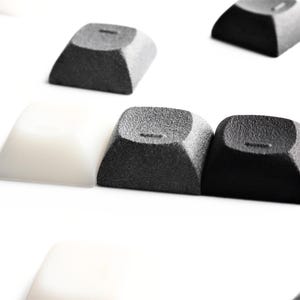 May include: A collection of individual mechanical keyboard keycaps arranged on a white surface. The set includes several matte black textured keycaps and a smooth, semi-translucent white keycap, each featuring a small horizontal dash indentation.