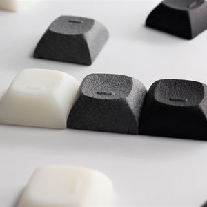 May include: Three black and white keycaps for a mechanical keyboard. The keycaps are made of a textured plastic and have a rounded shape.