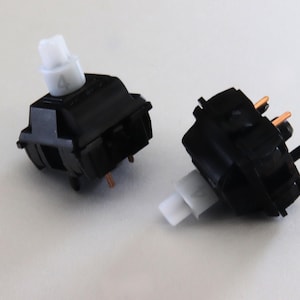 Könnte beinhalten: Zwei schwarze mechanische Tastaturschalter mit weißen Stielen. Auf einem Schalter ist die Zahl "4" auf dem Stiel aufgedruckt. Die Schalter haben kupferfarbene Stifte und sind vor einem weißen Hintergrund platziert.