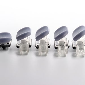 May include: Five gray and clear plastic mechanical keyboard switches are arranged in a row. Each switch has a gray, curved top and a clear plastic base. The bases have numbers 25, 30, 35, and 40 printed on them.