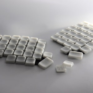 May include: Arrangement of small, square, off-white tiles. The tiles are slightly rounded and arranged in neat rows, with some tiles scattered. The lighting creates soft shadows, highlighting the texture and shape of the tiles.