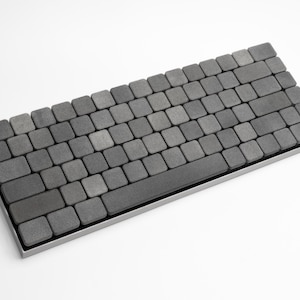 Puede incluir: Un teclado minimalista con teclas cuadradas grises. Las teclas están dispuestas en un diseño estándar, con una barra espaciadora larga. El teclado tiene un marco plateado y se encuentra sobre una superficie blanca. El diseño general es moderno y elegante.