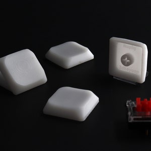 May include: A collection of white square keycaps and a red mechanical keyboard switch on a black surface. One keycap has a textured surface, and another has the text "Nephy 3dkeycap".