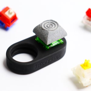 May include: A black 3dkeycap holder with a gray keycap and several mechanical keyboard switches. The keycap has a spiral design. The switches are red, blue, yellow, and white.