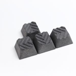 May include: Four dark gray, geometric keycaps with a textured chevron pattern on the top surface. The keycaps are arranged diagonally against a white background, showcasing their angular design and detail.