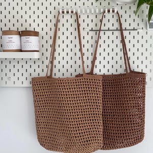 May include: Two brown crocheted tote bags hanging on a pegboard wall. The bags have a square mesh pattern and are made of a thick, textured yarn.