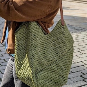 May include: A large, olive green crochet tote bag with a brown leather strap. The bag has a textured, woven appearance and is carried over the shoulder. The strap is attached to the bag with small rivets.