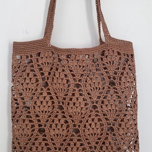 May include: A brown, open-weave crochet tote bag with two handles. The bag features a repeating floral pattern throughout the body and a solid border at the bottom. The bag is made of a soft, natural fiber.