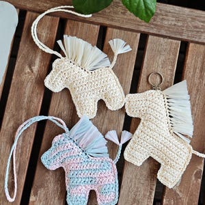 May include: Three crocheted horse ornaments in a top-down shot. Two are off-white with white fringe manes and tails, one with a keychain. The third is striped with pink and blue yarn, also with a white fringe mane and tail. All have a loop for hanging.