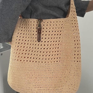 May include: A beige crochet tote bag with a long strap. The bag features a textured, open weave design and a brown leather strap closure. The bag is made of a light-colored yarn and is suitable for carrying various items.