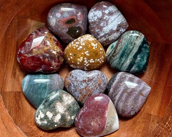 May include: A collection of heart-shaped polished stones in a wooden bowl. The stones display a variety of colors, including red, green, brown, and white. Some stones have speckled patterns, while others have swirling designs.