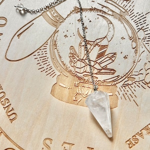 May include: A wooden pendulum board with a clear quartz crystal pendulum hanging from a silver chain. The board has the words "YES", "NO", "MAYBE", "REPHRASE", and "UNSOUL" inscribed on it.