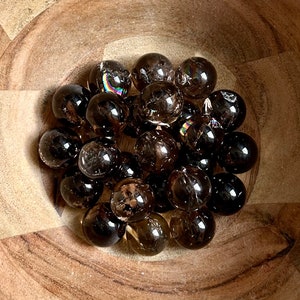 May include: A collection of smoky quartz crystal spheres in a wooden bowl. The spheres are various sizes and have a dark brown color with a slight iridescent sheen.