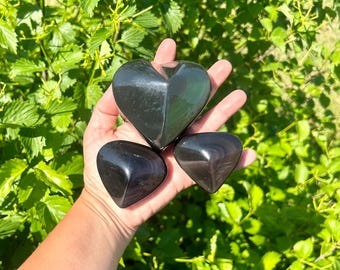 May include: Three polished obsidian heart-shaped stones. The largest heart is centered, with two smaller heart-shaped stones below. The stones are a deep, reflective black, held against a backdrop of green foliage.