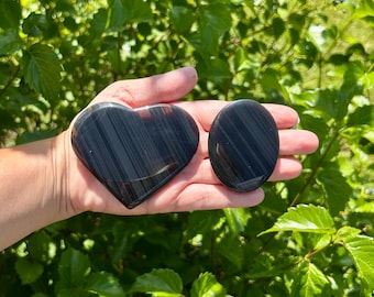 May include: Two polished obsidian stones, one heart-shaped and one oval, are held in a hand. The stones are black with subtle gray striations. The background is a lush green of leaves and foliage.