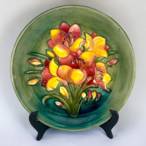 Könnte beinhalten: Ein grüner Keramikteller mit einem erhabenen Blumendesign. Die Blumen sind gelb, orange und rot. Der Teller steht auf einem schwarzen Ständer.