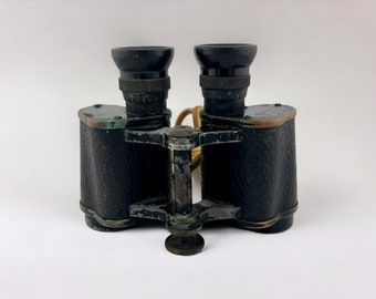Könnte beinhalten: Vintage-Fernglas aus schwarzem Leder mit Messingakzenten und einer abgenutzten grünen Oberfläche. Das Fernglas hat einen zentralen Fokusknopf und ein Band.