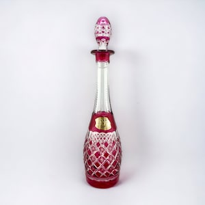 May include: A ruby-red glass decanter with a diamond-cut design and a matching stopper. The decanter has a long, slender neck and a rounded base. A gold label is attached. The stopper is intricately designed.
