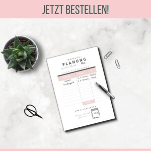 May include: A financial planning sheet with the text "MONATS-PLANUNG Mai" and "JETZT BESTELLEN!" is displayed on a white marble surface. A small succulent plant in a pot, a pair of black scissors, a pen, and two silver paper clips are also visible.