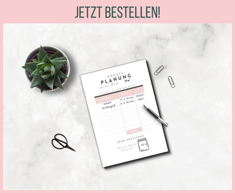 May include: A financial planning sheet with the text "MONATS-PLANUNG Mai" and "JETZT BESTELLEN!" is displayed on a white marble surface. A small succulent plant in a pot, a pair of black scissors, a pen, and two silver paper clips are also visible.
