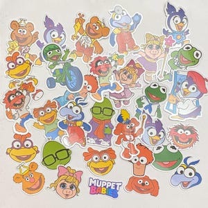 Peut inclure: Une collection d'autocollants colorés des Muppet Babies, représentant divers personnages comme Kermit la Grenouille, Miss Piggy et Gonzo. Les autocollants présentent les personnages dans différentes poses et tenues, avec le logo "Muppet Babies".