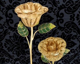May include: Two gold-colored rose sculptures with intricate detailing. The rose petals are open, and the stems have green and yellow leaves. The background is a dark, patterned fabric. These decorative pieces are likely for home decor.