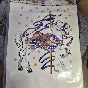 May include: Iron-on transfer featuring a carousel horse with patriotic colors and an eagle design. The horse is white with a blue, red, and white saddle and accents. The background is white with scattered orange stars. The package reads "Cache Junction".