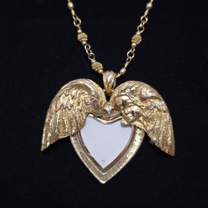 Puede incluir: Collar con colgante en forma de coraz&oacute;n dorado. El colgante presenta alas y rostros de &aacute;ngeles, con un centro blanco. Peque&ntilde;as piedras transparentes est&aacute;n engastadas en el dise&ntilde;o. El collar tiene una cadena delicada.