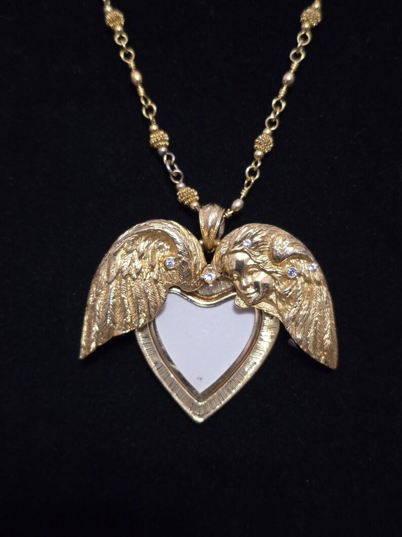 Puede incluir: Collar con colgante en forma de coraz&oacute;n dorado. El colgante presenta alas y rostros de &aacute;ngeles, con un centro blanco. Peque&ntilde;as piedras transparentes est&aacute;n engastadas en el dise&ntilde;o. El collar tiene una cadena delicada.