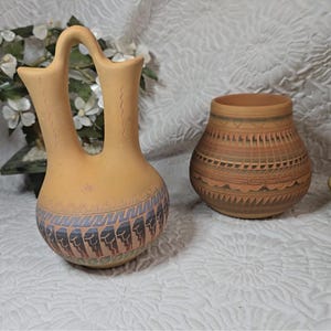 May include: Two handcrafted pottery vessels. One is a light tan double-spouted vase with a blue, black, and red geometric design. The other is a round, brown pot with intricate patterns in shades of brown and tan.