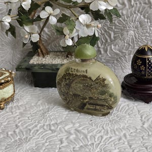May include: An oval-shaped, antique glass bottle with a green stopper. The bottle features a detailed sepia-toned landscape scene. A small, decorative jade tree with white flowers is in the background.