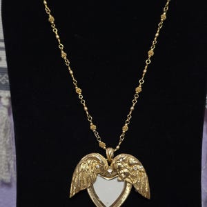 Puede incluir: Collar dorado con un colgante en forma de coraz&oacute;n con alas de &aacute;ngel y una peque&ntilde;a cara a cada lado. El colgante tiene un centro blanco y est&aacute; adornado con peque&ntilde;os cristales. La cadena est&aacute; hecha de peque&ntilde;as cuentas redondas.