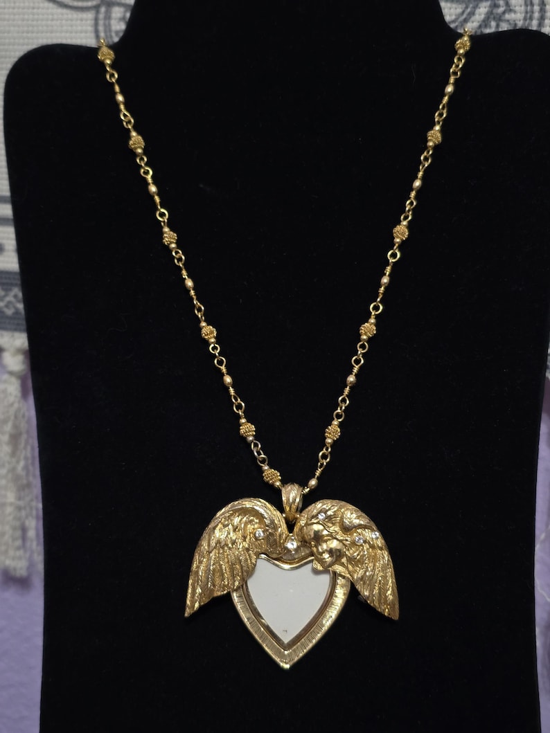 Puede incluir: Collar dorado con un colgante en forma de coraz&oacute;n con alas de &aacute;ngel y una peque&ntilde;a cara a cada lado. El colgante tiene un centro blanco y est&aacute; adornado con peque&ntilde;os cristales. La cadena est&aacute; hecha de peque&ntilde;as cuentas redondas.